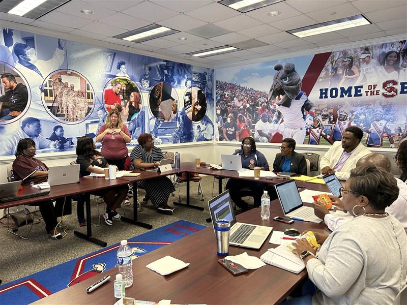 Shows an on campus workshop with participants sitting at tables with their laptops out listening to the speaker. 